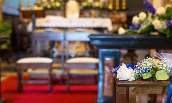 Floral arrangements decorate a wooden pew and altar; two kneelers are positioned in front of an altar adorned with white and blue flowers in a brightly lit church interior.