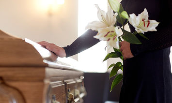 A person in black clothing holds white lilies and rests a hand on a wooden casket in a dimly lit room.