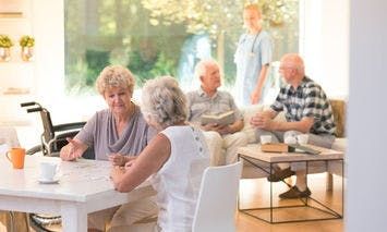 Elderly women sitting at a table solving a puzzle indoors, with a man and a healthcare worker conversing in the background near a window in a well-lit room.